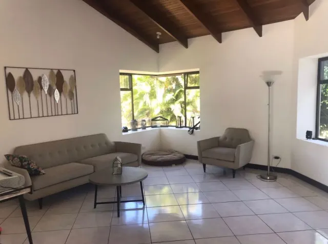 A living room featuring white walls, tiled floors, a grey sofa and armchair, a round coffee table, and windows showing outdoor greenery.