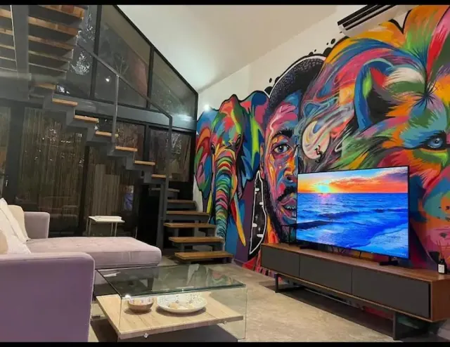 A living room featuring a large colorful wall mural, a television on a media console, a sofa, and an open metal staircase.