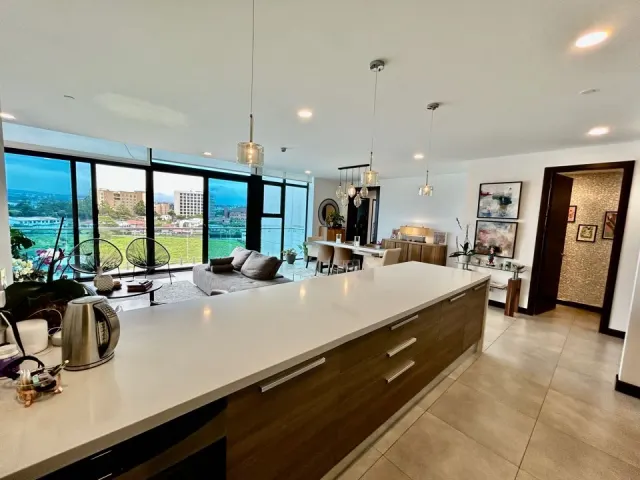 A long kitchen island with wood-veneer cabinets and a white countertop, overlooking an open-plan living area with floor-to-ceiling windows and outdoor views.