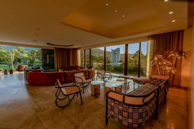 A spacious living area featuring a curved brown sofa, rattan armchairs, glass-topped coffee tables, and floor-to-ceiling windows with a view of greenery and buildings.