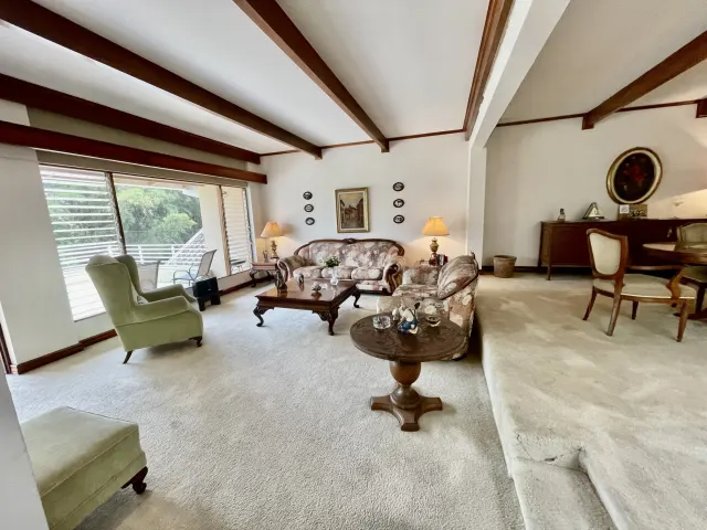 A carpeted living room featuring a patterned sofa set, wooden coffee tables, a green armchair, and large windows leading to a balcony.
