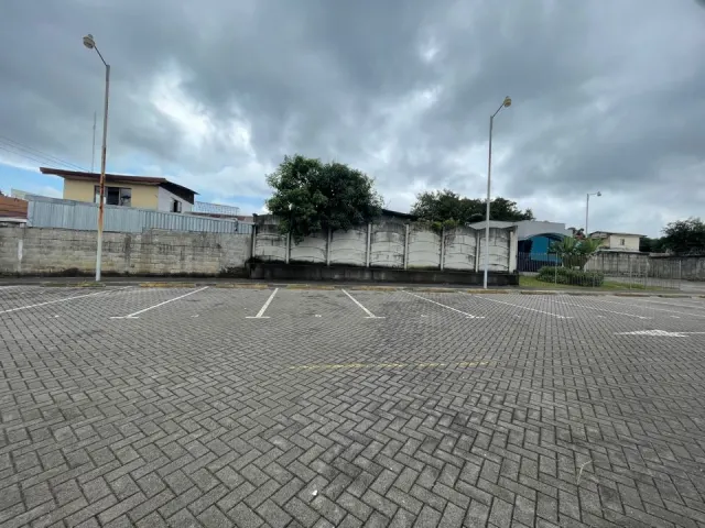 An outdoor parking lot paved with grey interlocking blocks features painted white parking stalls, light poles, and a perimeter wall under an overcast sky.
