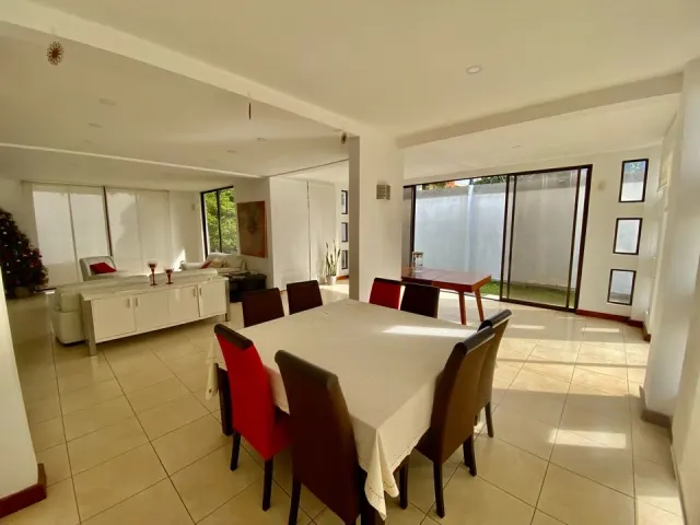 An open plan dining area with a square table and upholstered chairs, connecting to a bright living room with a white sofa and glass doors.