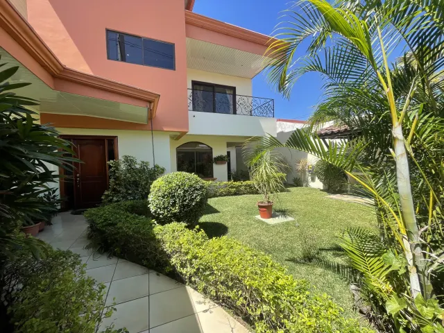 A green lawn and landscaped bushes sit in front of a two-story house with a peach exterior and a tiled walkway.