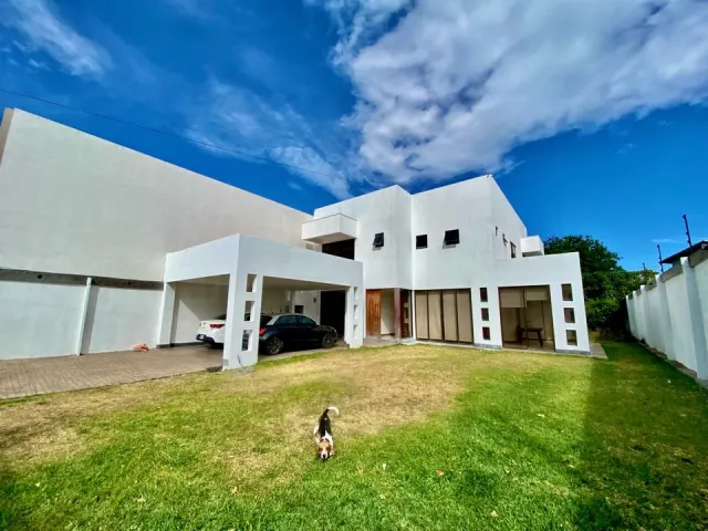 Two-story white exterior of a modern house featuring a covered carport with parked vehicles and a front lawn under a blue sky.