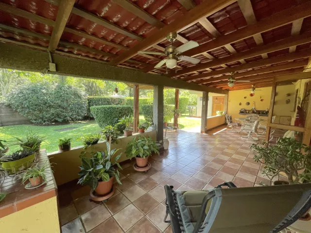 A tiled patio with an exposed wood ceiling, ceiling fans, and concrete pillars looking out onto a grassy garden with potted plants along the ledge.