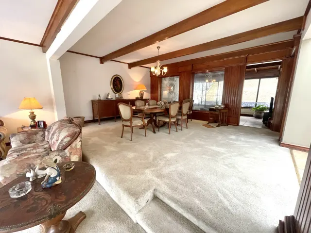 Carpeted living and dining area with exposed dark wooden beams on the ceiling, wood paneling, and an oval dining table with six chairs.