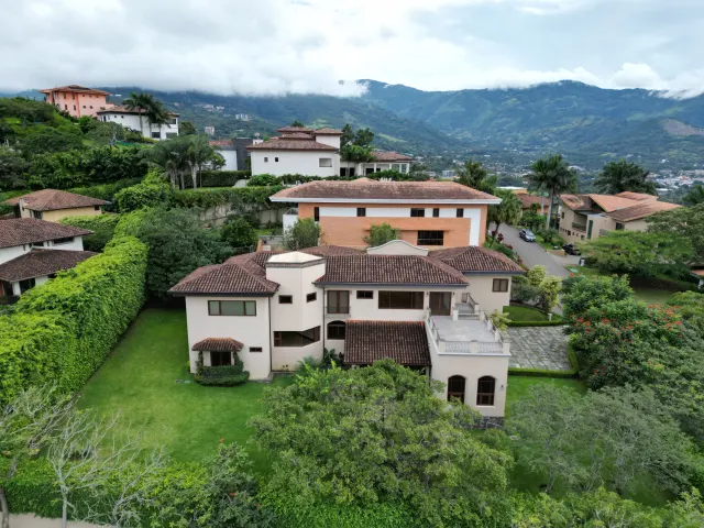 Aerial view of a multi-story house with a clay tile roof, white walls, and a large lawn, surrounded by trees and mountains.
