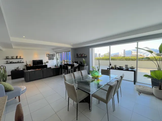 Living and dining space featuring a glass top dining table with six chairs, a dark sofa, and large windows leading to a balcony with city views.