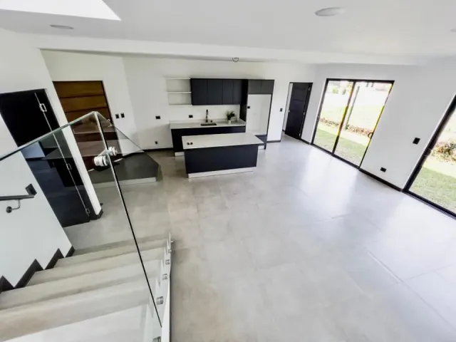 Open interior space showing a kitchen with dark cabinets and island, grey floor tiles, and a staircase with a glass railing.