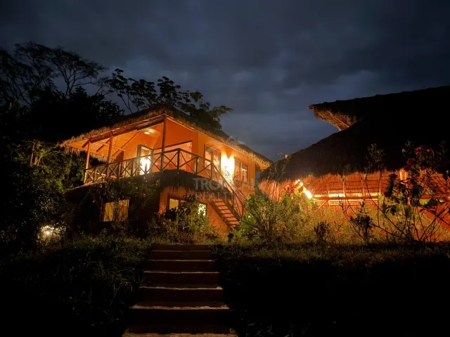 Exterior view at night of a two-story house with a thatched roof, lit windows, a wooden balcony, and stone steps leading up from the foreground.