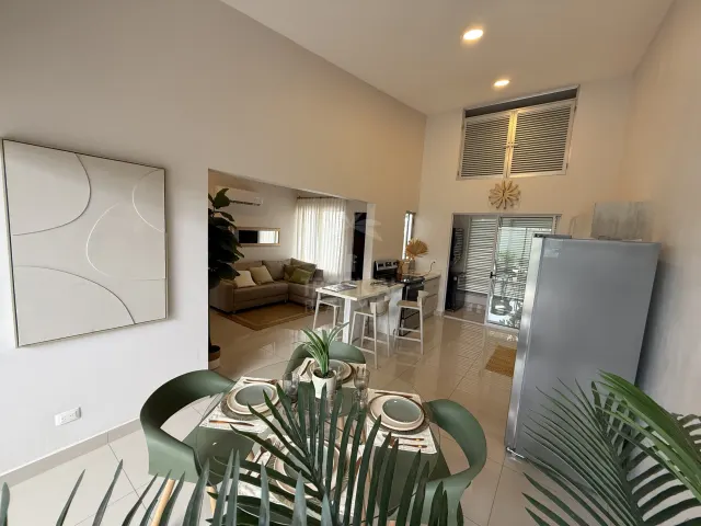 An interior view showing a dining table in the foreground, a kitchen with a white island, and an adjacent living room with a sofa.