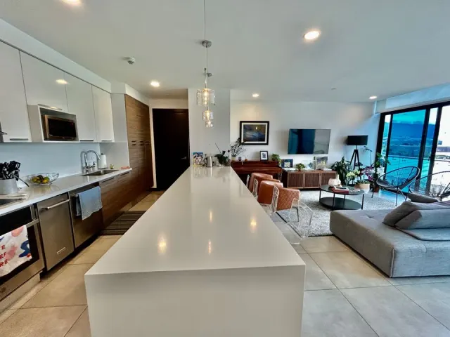 Open plan living room and kitchen featuring a long white kitchen island, glass pendant lights, and a grey sectional sofa.