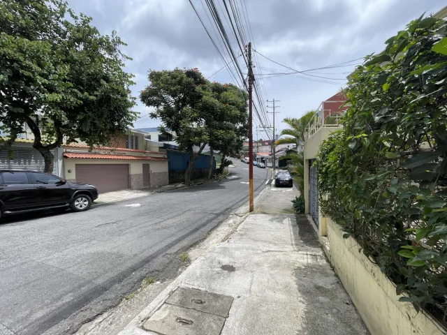 View of a paved street with a sidewalk, overhead utility lines, and parked vehicles under an overcast sky.