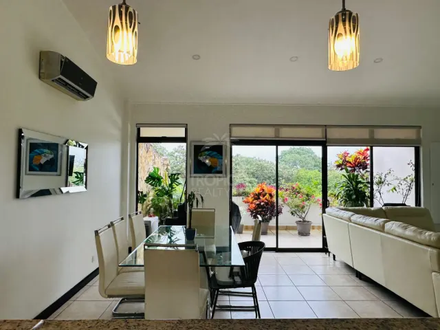 Living and dining space featuring a glass-topped dining table, white chairs, two pendant lights, and sliding glass doors opening to a planted balcony.