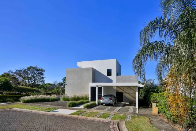Exterior view of a modern two-story concrete house featuring a covered carport with a parked car, surrounded by manicured grass and trees under a clear blue sky.