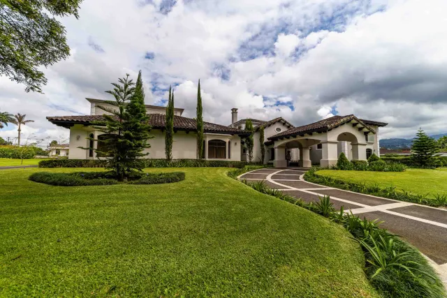 Exterior view of a light-colored home with a tiled roof, curved driveway, and manicured lawn under a cloudy sky.