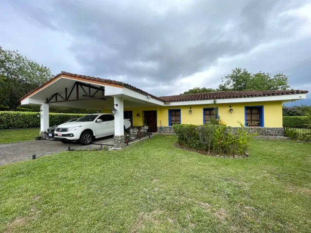 One-story yellow house with a tile roof, blue window frames, stone foundation, and a covered carport holding two vehicles.