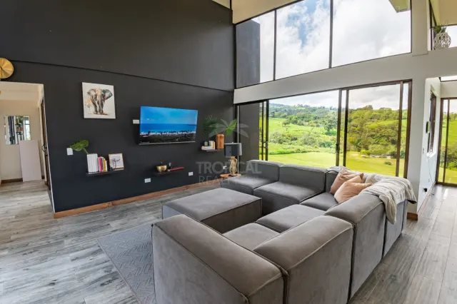 High ceiling living room featuring a dark accent wall, wall-mounted television, large sectional sofa, and floor-to-ceiling windows showing green hills.