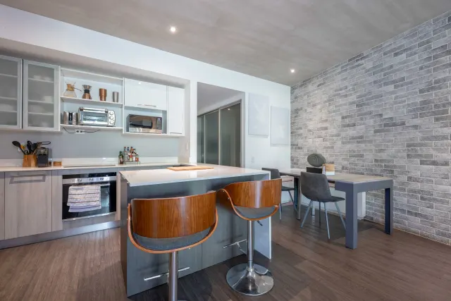 Kitchen with white upper cabinets, stainless steel appliances, and an island with two wooden bar stools. Next to it is a dining table with grey chairs against a grey brick wall.