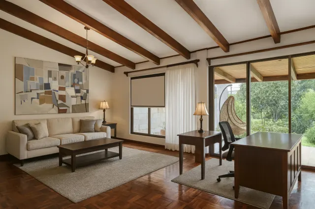 Combination living room and workspace featuring a beige sofa, dark wood desks, wood parquet flooring, and exposed ceiling beams. Sliding glass doors look out to greenery.