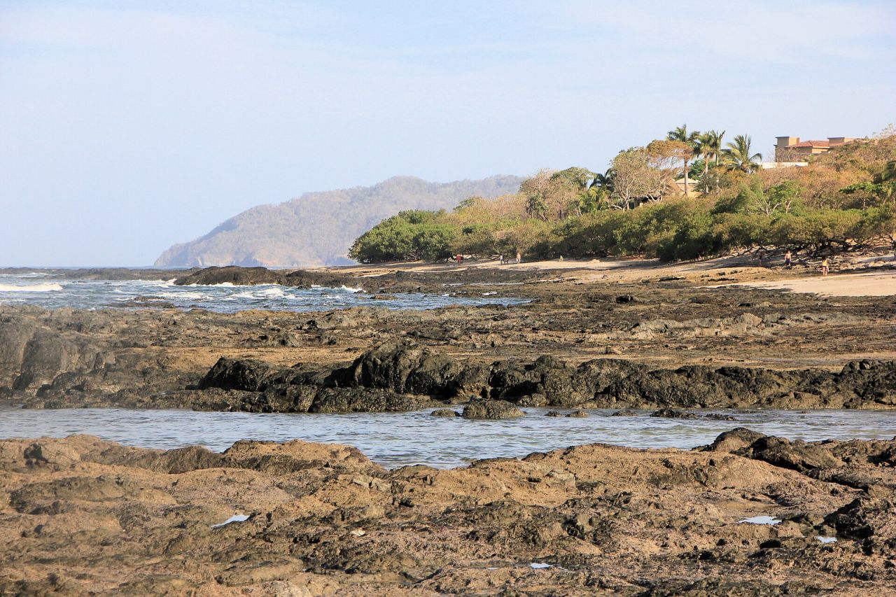 Rocky tidal shelf and pale sand at Playa Langosta, Guanacaste, with beachfront homes among the palms